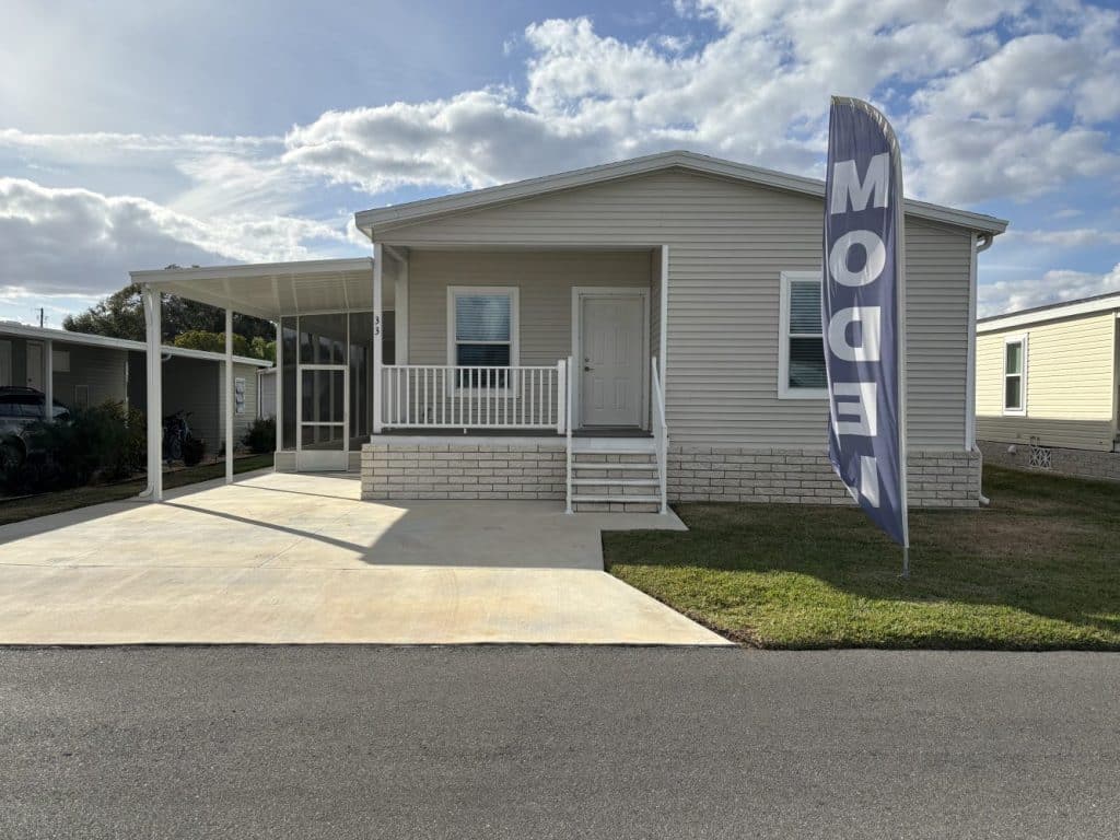 Single-story manufactured home with a covered carport, front porch, and a large "MODEL" flag on the lawn, under a partly cloudy sky.