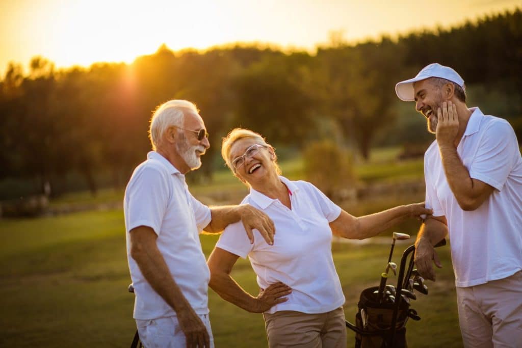 Three older adults in white golf attire laugh together beside golf bags on a golf course at sunset.