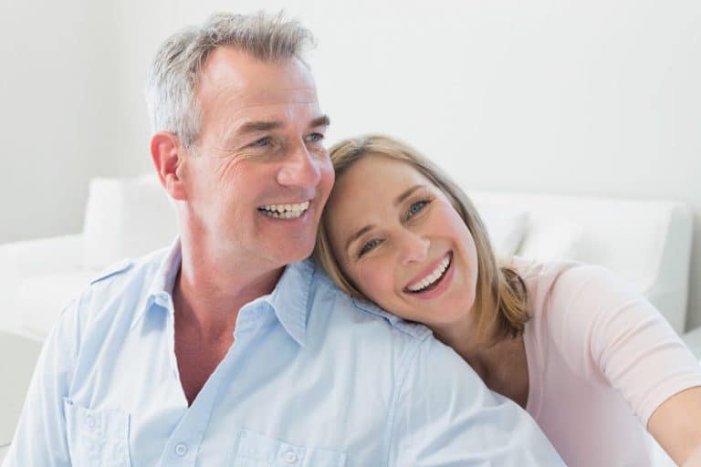 Smiling middle-aged couple sitting together indoors, with the woman resting her head on the man's shoulder.