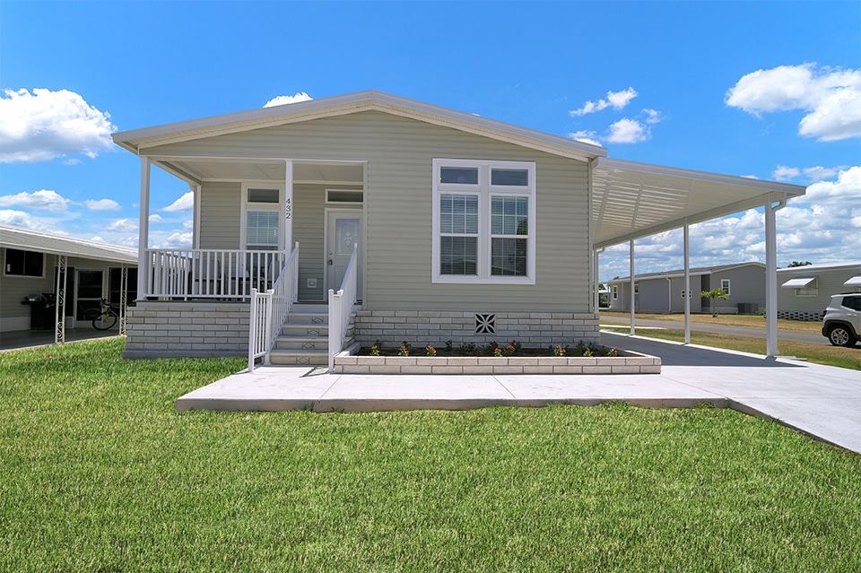 A beige manufactured home with a covered front porch, large front windows, and a driveway, set on a manicured lawn under a blue sky with scattered clouds.