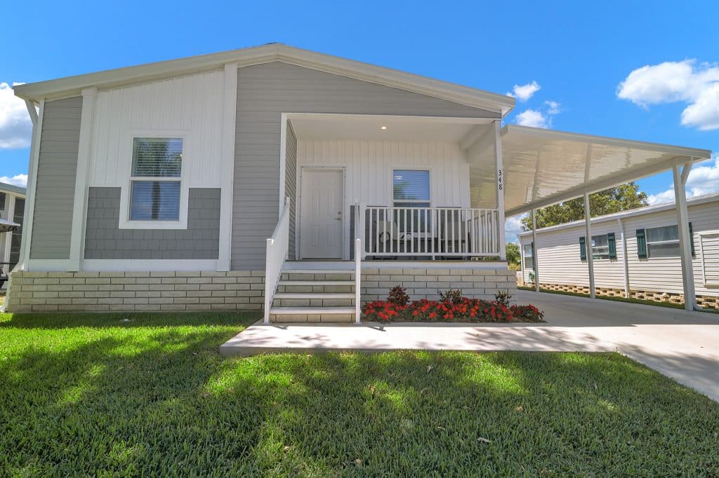 Single-story manufactured home with a front porch, steps, railing, flower bed, and carport, set on a lawn under a clear blue sky.