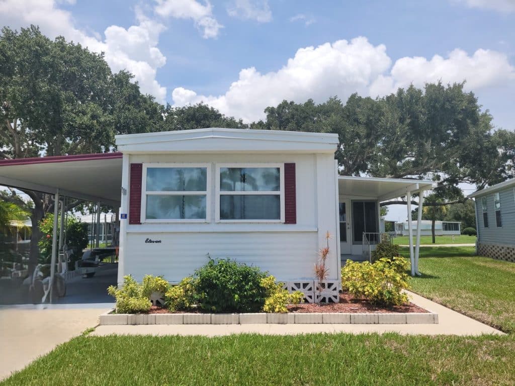 A white mobile home with red shutters, small front garden, and covered carport, set on a sunny day with trees and grass surrounding the property.