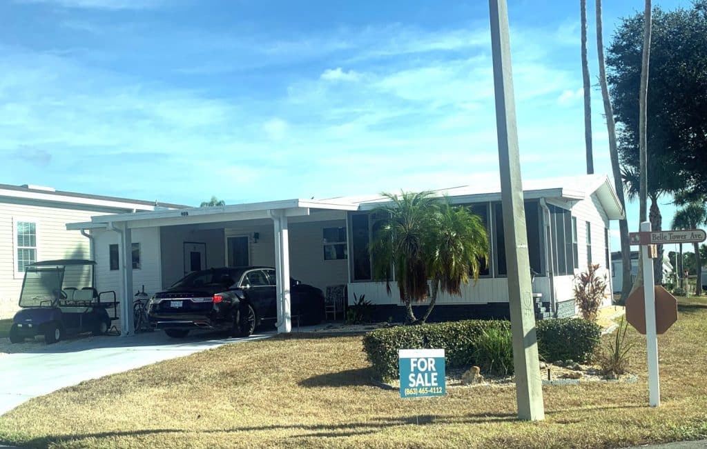 A single-story manufactured home with a car and a golf cart parked in the driveway, a palm tree, and a "For Sale" sign in the yard.