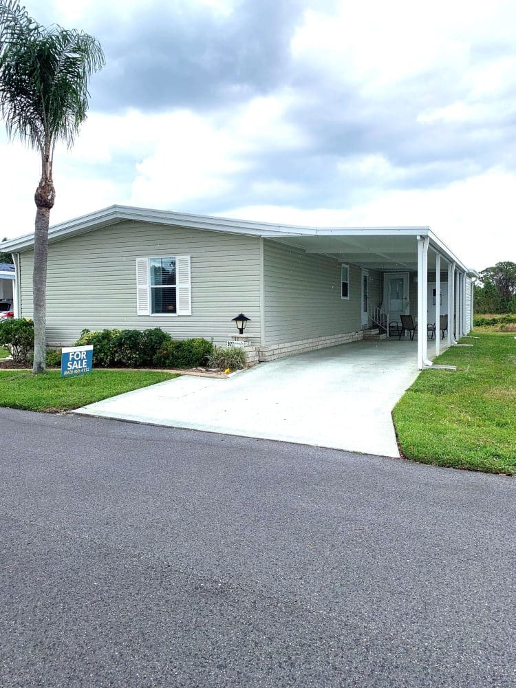 Single-story manufactured home with covered carport, small front porch, a "For Sale" sign in the yard, and a palm tree near the driveway.