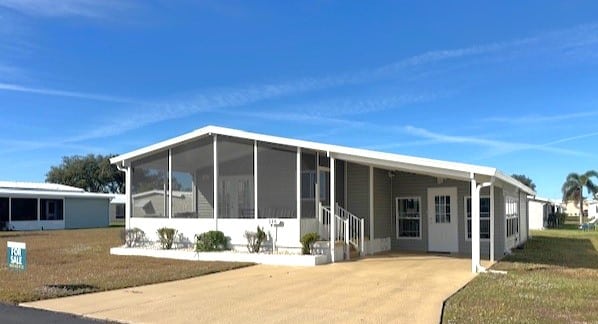 Single-story manufactured home with a screened front porch, driveway, and "For Sale" sign on a grassy lot under a clear blue sky.