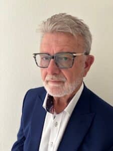 An older man with gray hair, a beard, and glasses wearing a white shirt and dark blue blazer stands against a plain light-colored wall.