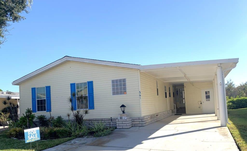 Single-story yellow manufactured home with blue shutters, a covered carport, front steps, and a "For Sale" sign in the yard under a clear blue sky.