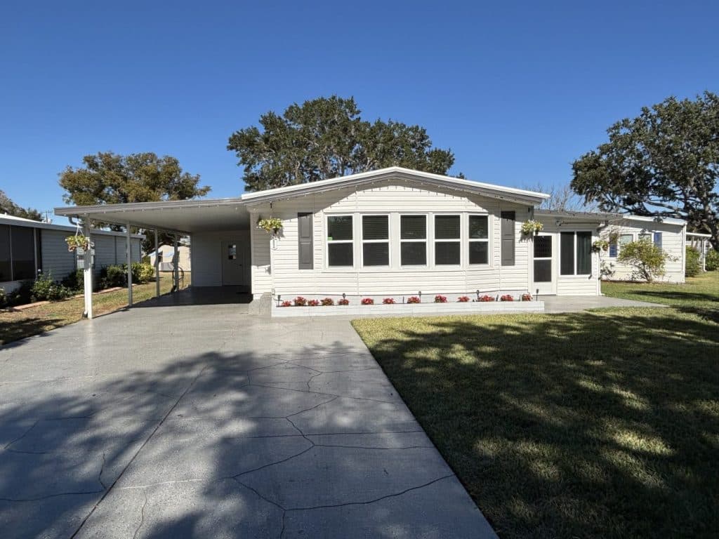 Single-story manufactured home with a covered carport, large front windows, hanging plants, and a flower bed, set on a sunny day with a clear blue sky.