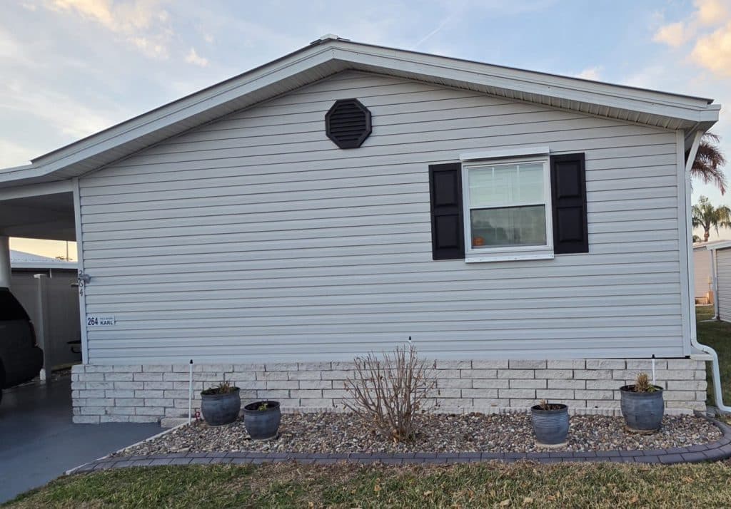 The side of a light gray manufactured home with one window, black shutters, five potted plants, a bare bush, and a driveway partially visible on the left.