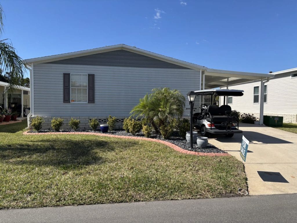 Single-story manufactured home with light gray siding, a small garden, and a driveway with a golf cart parked under a carport on a sunny day.