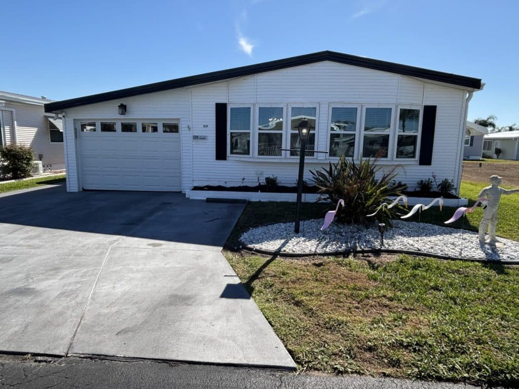 Single-story white house with black trim, two-car garage, concrete driveway, small landscaped yard, and a lawn decoration with pink flamingos and a statue.