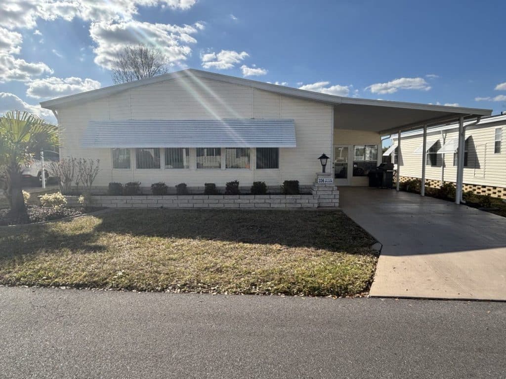 Single-story manufactured home with a covered carport, front lawn, and bushes under windows, photographed on a sunny day with a blue sky and scattered clouds.