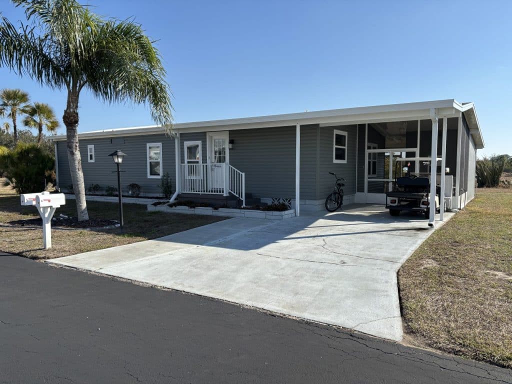 Gray single-story manufactured home with a covered carport, front porch, mailbox, palm tree, bicycle, and golf cart on a concrete driveway under a clear blue sky.