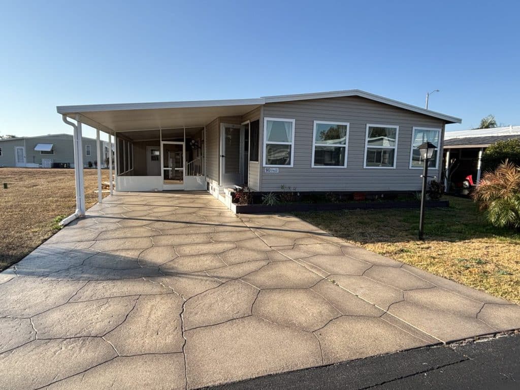 Single-story manufactured home with beige siding, multiple front windows, a covered carport on the left, concrete driveway, and small landscaped area in front, under a clear blue sky.