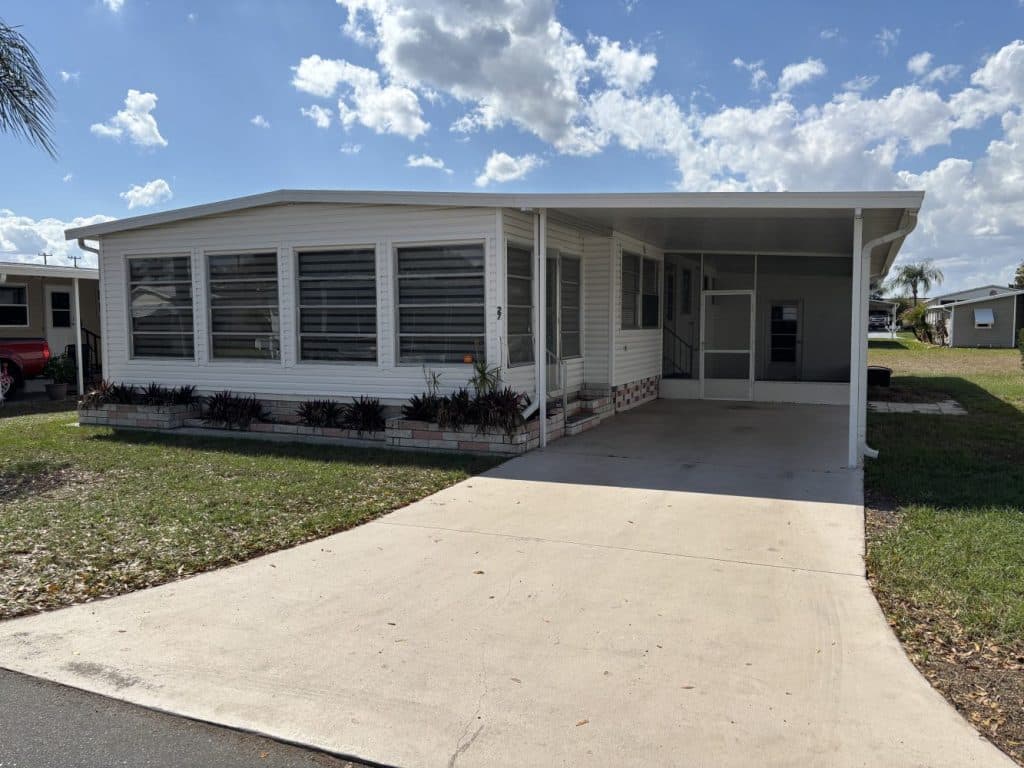 Single-story white manufactured home with a covered carport, front sunroom, and a concrete driveway under a partly cloudy sky.