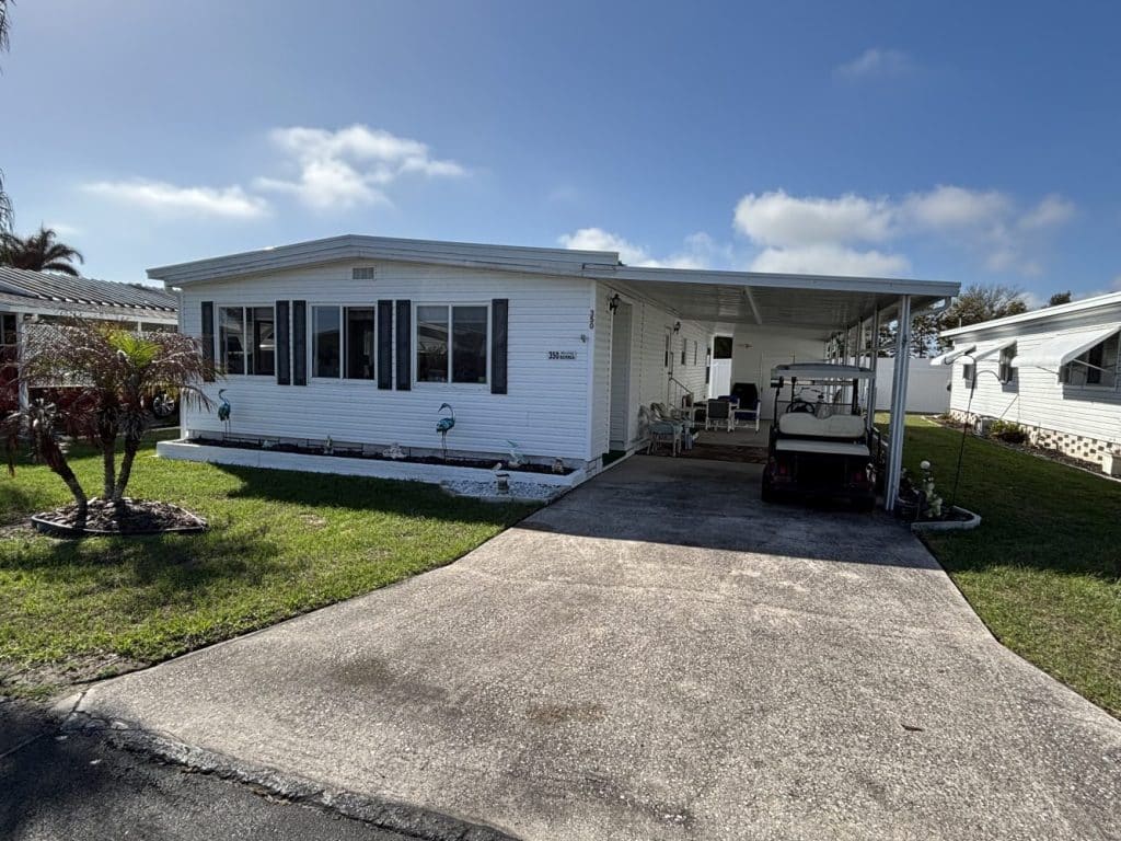 A white mobile home with a carport, golf cart, small lawn, and a palm tree in front, under a blue sky with some clouds.