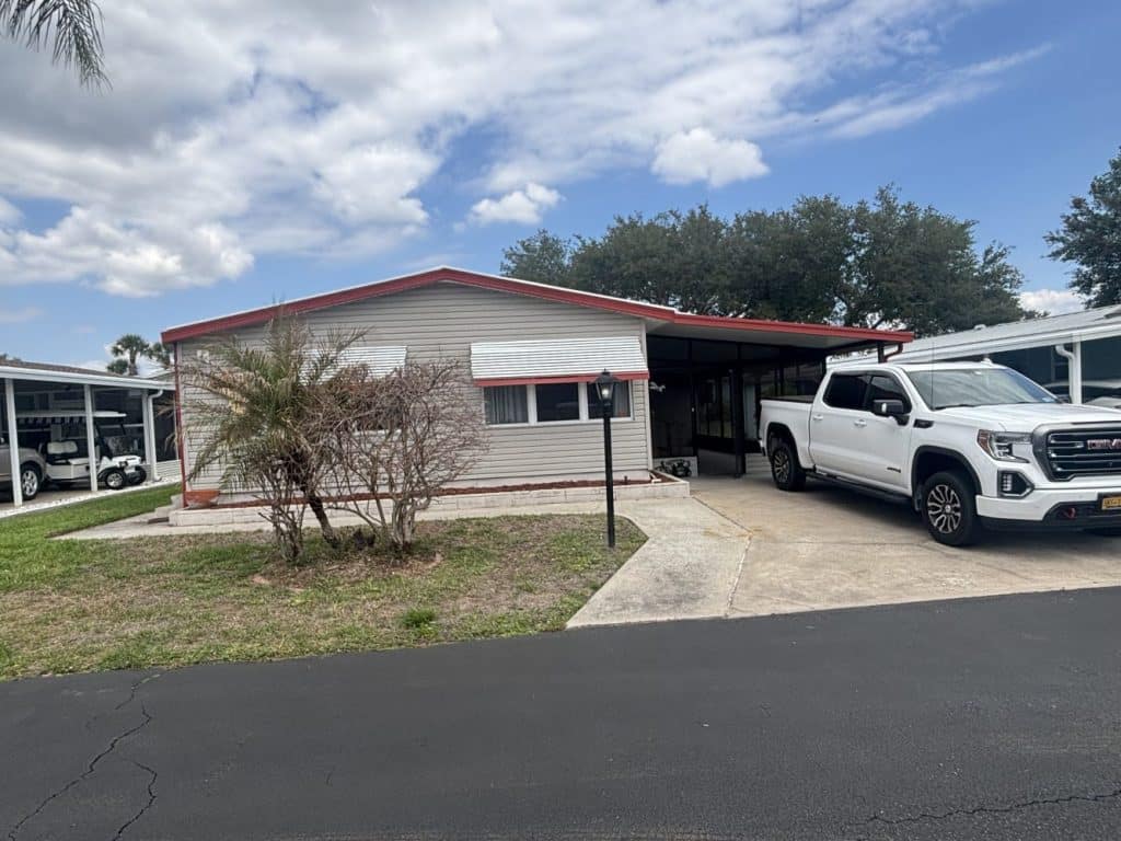 A one-story mobile home with a carport, white truck parked outside, and cloudy sky overhead.