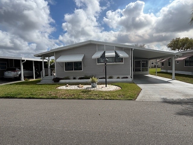 Single-story manufactured home with white siding, awnings over windows, a small landscaped front yard, and a covered driveway on a paved street under a partly cloudy sky.