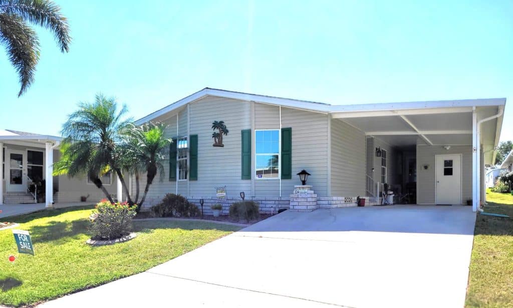 Single-story beige manufactured home with green shutters, carport, palm trees, and a "For Sale" sign on the lawn.
