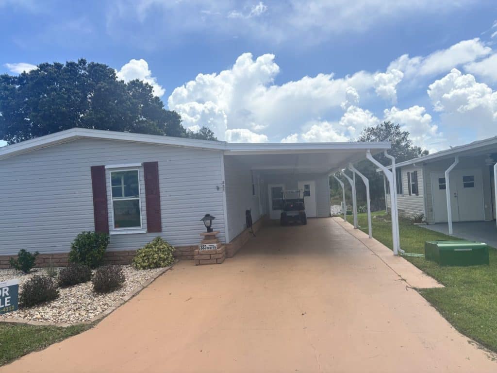 Single-story mobile home with carport, red shutters, and landscaped front yard under a partly cloudy sky.