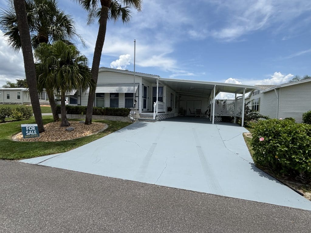 White manufactured home with a large driveway, carport, palm trees, and a "For Sale" sign on the lawn.