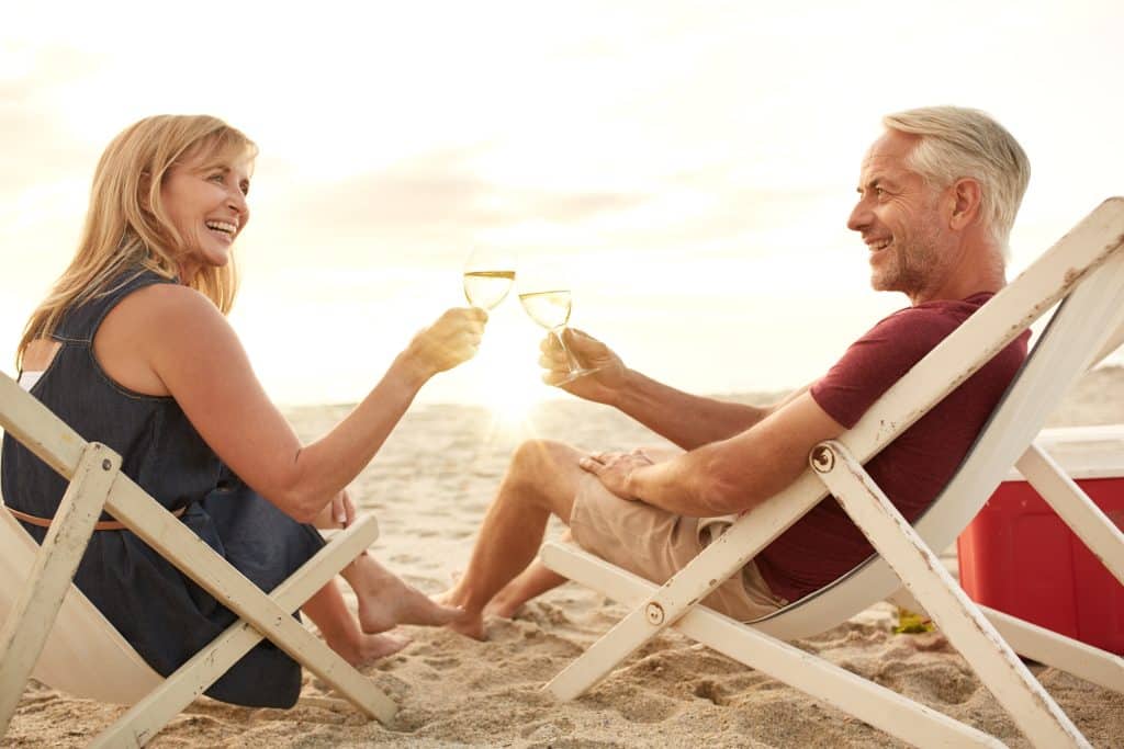 Smiling couple in deck chairs on the beach, toasting with wine glasses, enjoying the sunset.