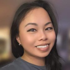 A woman with long dark hair and a gray shirt smiles at the camera against a softly blurred indoor background.