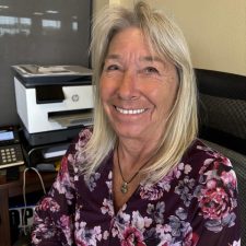 A woman with long blonde hair wearing a floral blouse sits and smiles at a desk with a printer, phone, and office supplies in the background.